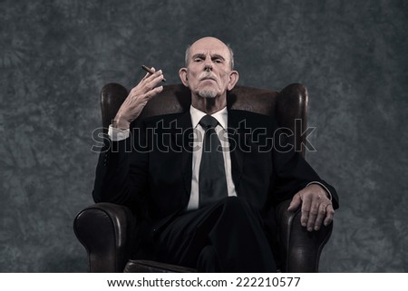 Cigar smoking senior businessman with gray beard wearing dark suit and tie. Sitting in leather chair. Against grey wall.