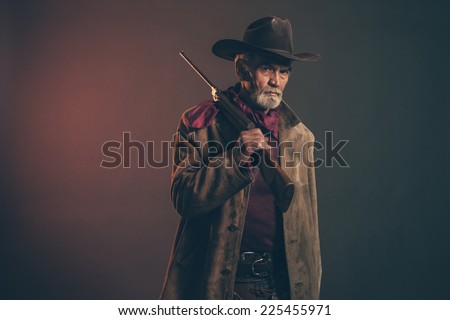Old rough western cowboy with gray beard and brown hat holding rifle. Low key studio shot.