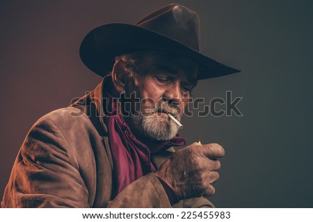 Old rough western cowboy with gray beard and brown hat lighting a cigarette. Low key studio shot.