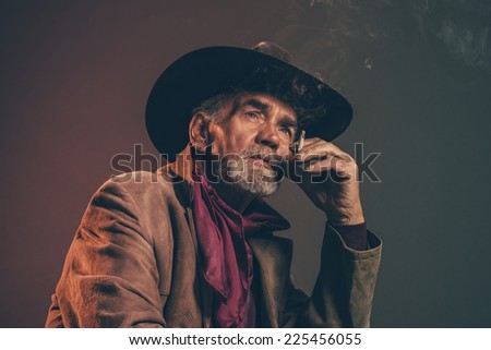 Old rough western cowboy with gray beard and brown hat smoking a cigarette. Low key studio shot.