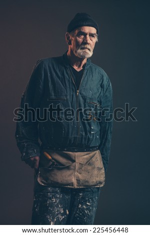 Senior carpenter with gray hair and beard wearing black hat with green overalls. Low key studio shot.
