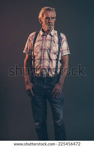 Characteristic senior man with gray hair and beard wearing checkered shirt with braces and blue jeans. Low key studio shot.