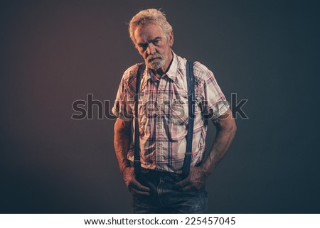 Characteristic senior man with gray hair and beard wearing checkered shirt with braces and blue jeans. Low key studio shot.