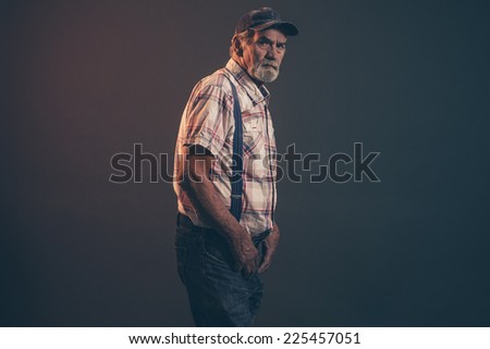 Characteristic senior man with gray hair and beard wearing blue cap with braces and jeans. Low key studio shot.