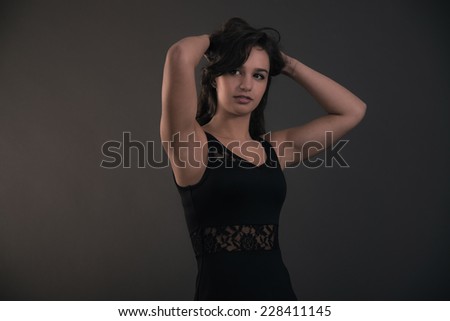 Cute brunette teenage girl with hands in hair wearing black dress. Studio fashion shot.