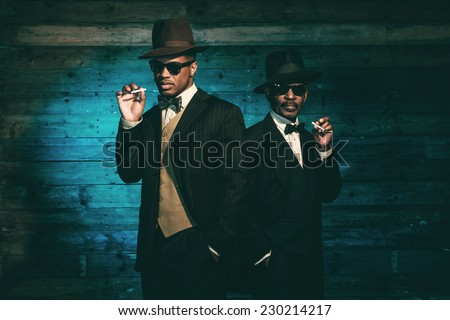 Two vintage african american gangsters smoking cigarette in front of old wooden wall. Wearing black sunglasses with suit and hat.