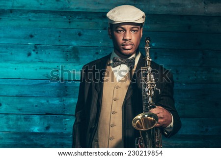Vintage african american jazz musician with saxophone in front of old wooden wall. Wearing suit and cap.