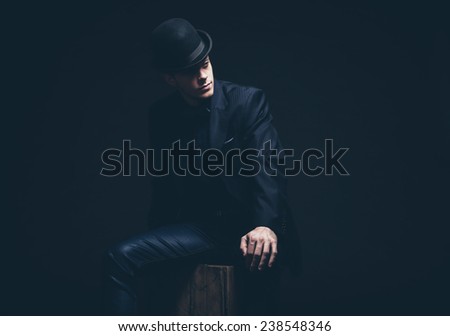 Retro fashion man with blue shirt, jacket, jeans and bow tie. Wearing black hat. Sitting on vintage wooden box. Studio shot against black.