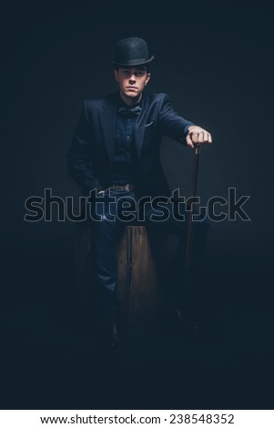 Retro fashion man with blue shirt, jacket, jeans and bow tie. Wearing black hat. Holding cane. Sitting on vintage wooden box. Studio shot against black.