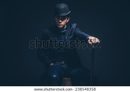 Retro fashion man with blue shirt, jacket, jeans and bow tie. Wearing black hat. Holding cane. Sitting on vintage wooden box. Studio shot against black.