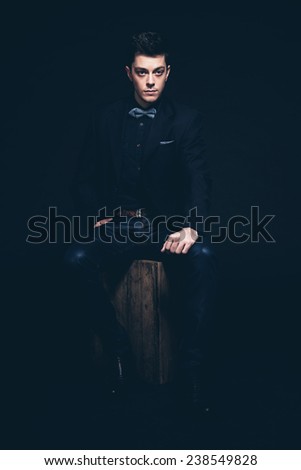 Retro fashion man with blue shirt, jacket, jeans and bow tie. Sitting on vintage wooden box. Short dark hair. Studio shot against black.