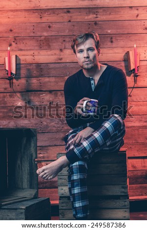 Man with blonde hair and beard wearing winter sleepwear. Sitting on wooden box inside cabin.