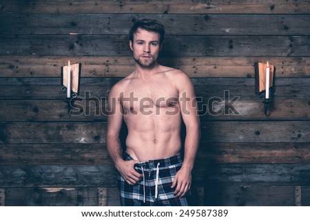 Man with blonde hair and bare chest wearing blue flannel pants. Standing against wooden wall inside wooden cabin.
