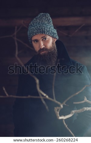 Close up Handsome Young Man with Long Goatee, Wearing Winter Fashion Behind Dry Twigs and Smoke, Looking at the Camera.