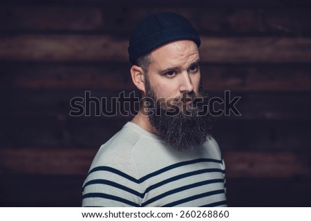 Close up Good Looking Man With Long Goatee Wearing Stripe Shirt and Bonnet on a Wooden Wall Background.