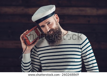 Close up Stylish Handsome Man with Long Beard in Sailor Attire, Holding a Vintage Radio While Looking at the Camera.