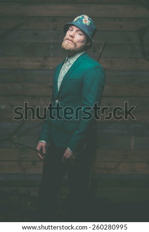 Middle Age Man with Goatee Posing in Green Formal Wear with Floral Hat on Vintage Wooden Wall Background While Looking at the Camera.
