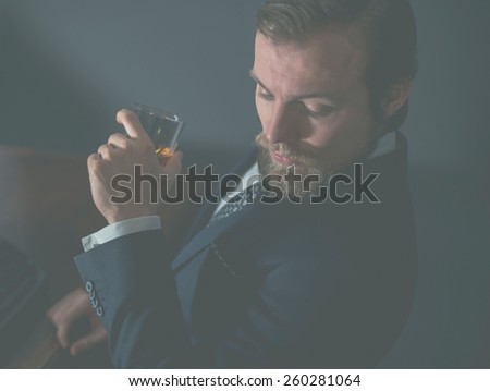 Close up of a stylish handsome bearded man enjoying a brandy or whiskey while looking thoughtfully downwards, aged effect
