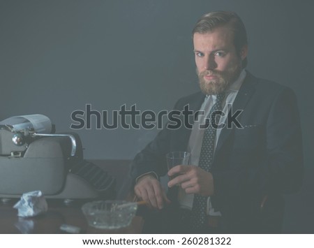 Moody portrait of a stylish vintage businessman sitting relaxing at his desk in the office in front of an old-fashioned manual typewriter with a drink in his hand