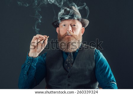 Close up Stylish Adult Man with Long Beard, in Denim Long Sleeves Shirt with Gray Vest and Cap, Smoking a Cigarette While Looking at the Camera. Isolated on a Gray Background.