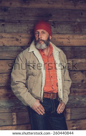 Close up Portrait of a Senior Goatee Man in Trendy Clothing with Red Bonnet Leaning on the Wooden Wall While Looking at the Camera.