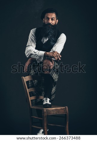 Portrait of a Bearded Stylish Man Standing with One Foot on a Chair and Cigarette on his Fingers While Smiling at the Camera on a Black Background.
