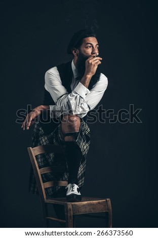 Thoughtful Scotsman with a bushy beard wearing his plaid kilt standing with one foot on a chair smoking and staring off into the distance