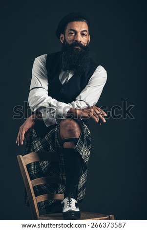 Serious thoughtful bearded Scotsman in a kilt standing with one foot up on a chair smoking a cigarette over a dark background
