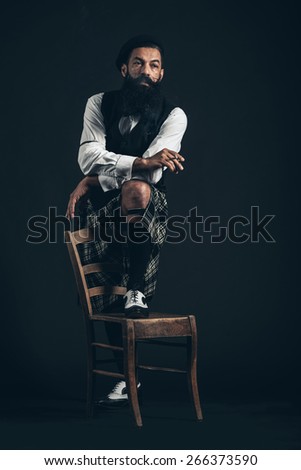 Portrait of a Serious Man with Long Beard Looking Afar, Holding a Cigarette with One Foot on Chair. Captured in Studio with Black Background.