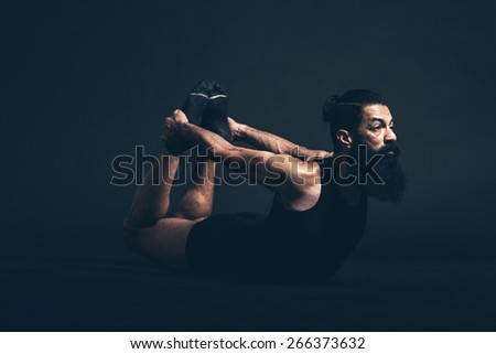 Athletic Guy with Long Beard and Mustache Doing Dhanurasana Bow Yoga Pose While Facing to the Right on a Black Background.