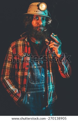 Male Miner Wearing Lit Safety Helmet Lamp, Overalls and Plaid Shirt Standing with Hand in Pocket and Smoking Cigarette