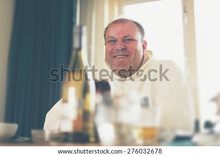 Happy overweight middle-aged man in casual clothes sitting at a dining table viewed across bottles and dinnerware, low angle