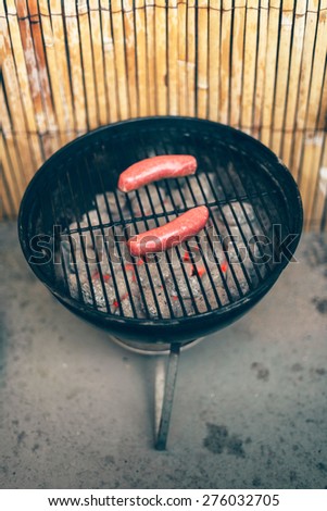 Two beef sausages cooking over the hot coals on an outdoor grill during a summer barbecue in the backyard with a bamboo fence visible behind