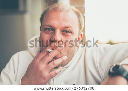 Balding middle-aged man sitting smoking indoors puffing on his cigarette with obvious enjoyment, close up head and shoulders portrait