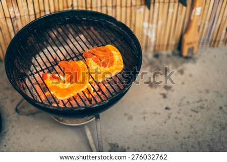 Wedges of fresh pumpkin grilling over the hot coals on a BBQ outdoors in the summer sunshine alongside a bamboo fence in the backyard