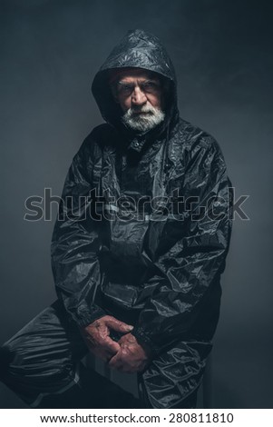 Portrait of a Serious Bearded Senior Man in Black Raincoat, Sitting on a Stool and Looking at the Camera Against Black Background.