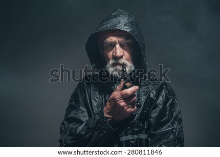 Portrait of a Reflective Bearded Old Guy in Black Rain Jacket, Smoking a Cigarette Using a Pipe. Captured in Studio.