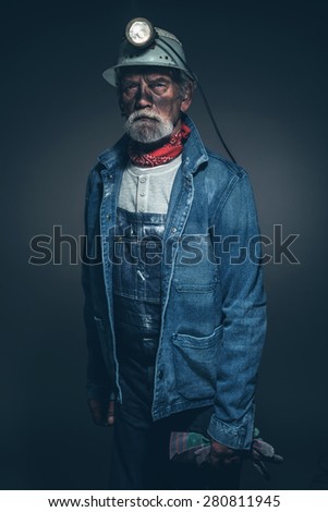 Portrait of a Dirty Senior Male Gold Miner, Wearing Denim Jacket and Helmet with Light, Holding his Gloves and Looking at the Camera Against Gray Gradient Background.