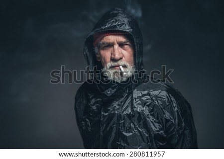 Portrait of a Serious Old Guy with Facial Hair in Rain Slicker Outfit, Smoking a Cigarette While Looking Straight at the Camera on a Black Background.