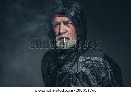Close up Bearded Adult Guy in Waterproof Jacket, Smoking a Cigarette in a Pensive Facial Expression. Captured in Studio with Black Background.