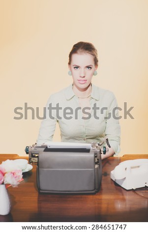 Vintage 1950 blonde secretary woman sitting behind desk working on typewriter. Looking towards camera. High angle view.