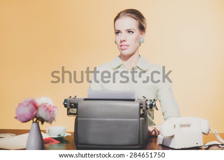 Pensive vintage 1950 blonde secretary woman sitting behind desk working on typewriter.