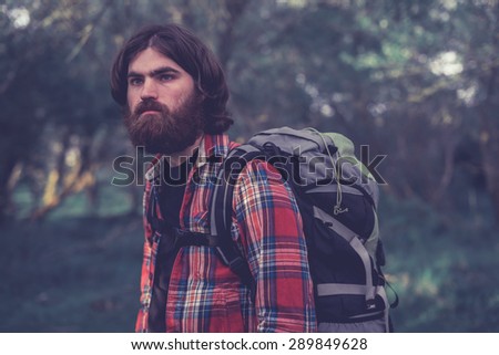 Handsome bearded male backpacker with a heavy backpack on his shoulders standing looking into the distance with a thoughtful expression in the shade of some trees