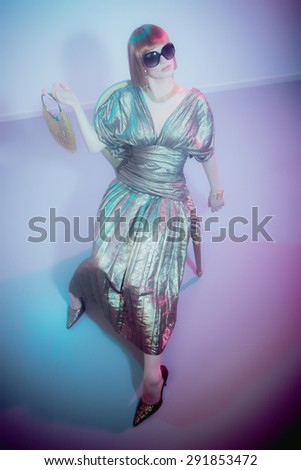 High Angle Full Length View of Glamorous Woman with Red Hair Wearing Sunglasses and Shiny Retro Gown Sitting in Chair Looking Bored and Holding Fringed Purse in Smoky Disco Night Club Setting