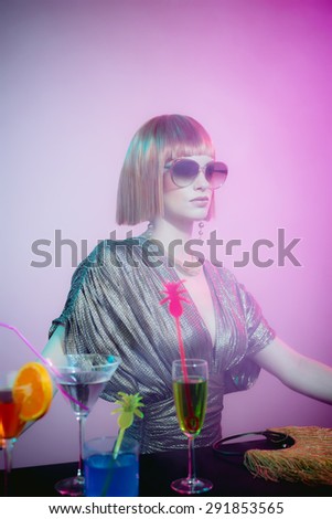 Glamorous and Sophisticated Woman with Red Hair Wearing Shiny Retro Gown and Sunglasses Leaning on Bar Covered in Cocktails in Smoky Disco Night Club Bar