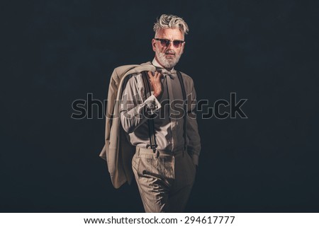 Elegant Senior Bearded Man in Formal Outfit with Sunglasses, Holding his Coat Over his Shoulder Against Black Background.