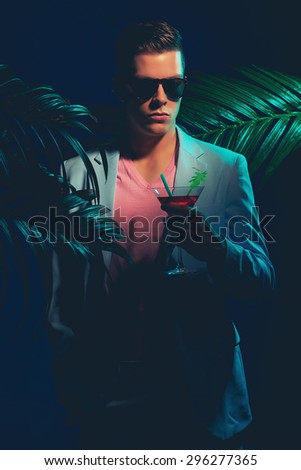Half Body Shot of a Gorgeous Young Man Wearing Formal Suit, Holding a Cocktail Drink While Standing Between Palm Leaves on a Dim Light.