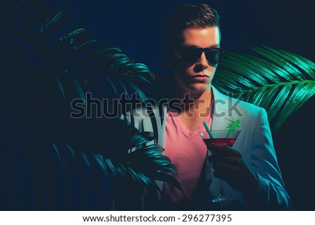 Close up Gorgeous Young Man Wearing Formal Suit, Holding a Cocktail Drink While Standing Between Palm Leaves on a Dim Light.