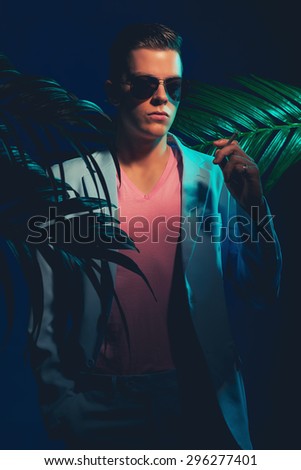 Half Body Shot of a Handsome Young Man Wearing Formal Suit and Sunglasses, Standing Between Palm Leaves While Holding a Cigarette and Looking Into the Distance on a Dimmer Light.
