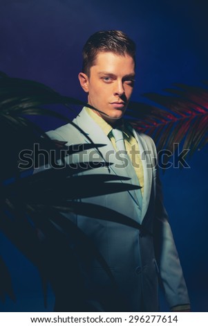 Half Body Shot of a Handsome Man in Formal Wear Standing Between Palm Leaves and Looking at the Camera Against Dark Blue Violet Background.
